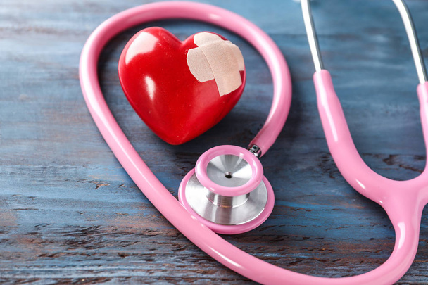 Medical stethoscope and red heart with plaster on wooden background, closeup. Cardiology concept - Photo, Image