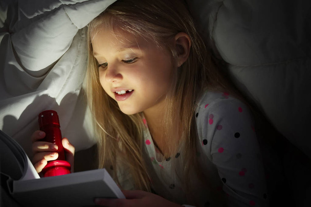 Cute little girl with flashlight reading book in bed under blanket - Фото, изображение