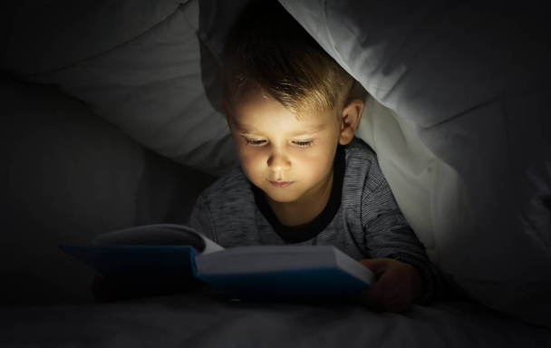 Cute little boy reading book in bed under blanket - Foto, afbeelding
