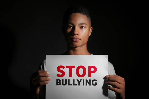 Un adolescent afro-américain tenant une feuille de papier avec du texte STOP BULLYING sur fond sombre
 - Photo, image