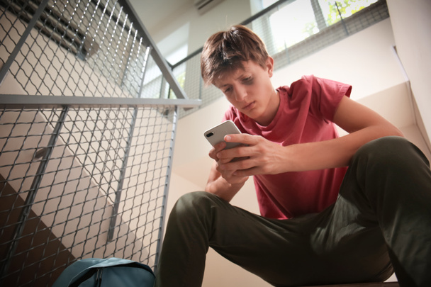 Sad teenage boy with mobile phone sitting on stairs. Bullying at school - Fotografie, Obrázek