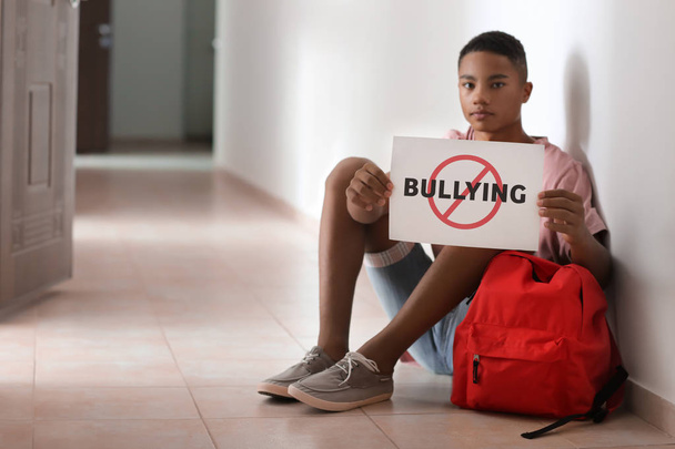 Adolescente afroamericano sosteniendo hoja de papel con palabra BULLYING mientras está sentado en el suelo en la escuela
 - Foto, Imagen