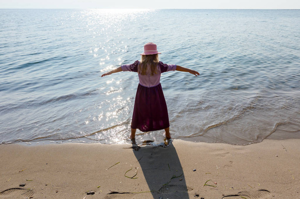 Молодая женщина, ребенок в шляпе и милое летнее платье делает жест с поднятыми руками на берегу моря, в мелководье
. - Фото, изображение