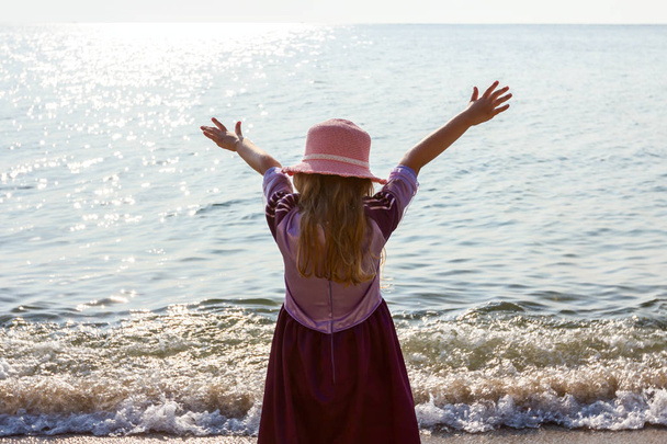 Νεαρή γυναίκα, το παιδί σε καπέλο και χαριτωμένο καλοκαιρινό φόρεμα κάνοντας χειρονομία με υψωμένα τα χέρια στην ακτή της θάλασσας, σε ρηχά νερά. - Φωτογραφία, εικόνα