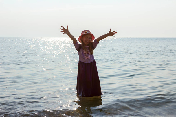 Fiatal nő, gyermek a kalapot, és aranyos nyári ruha teszi gesztus, a tenger partján emelt kezével sekély vízben. - Fotó, kép