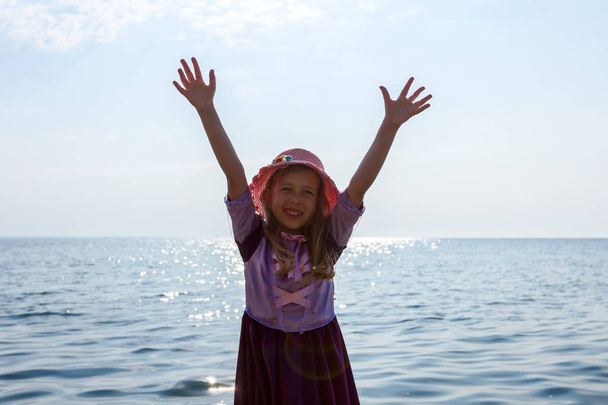 Young woman, child in hat and cute summer dress is making gesture with raised hands on the sea shore, in shallow water. - Photo, Image