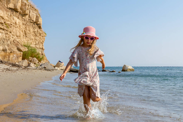 Young woman, child in hat and cute summer dress is running and splashing in shallow water, the sea shore. - Photo, Image