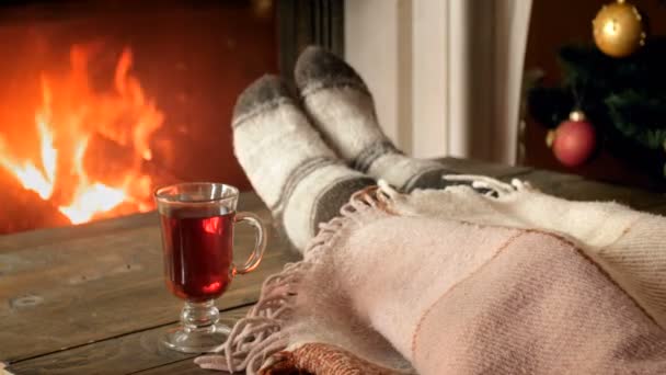 Closeup 4k video of female feet under blanket and cup of tea on wooden table at burning fireplace - Footage, Video