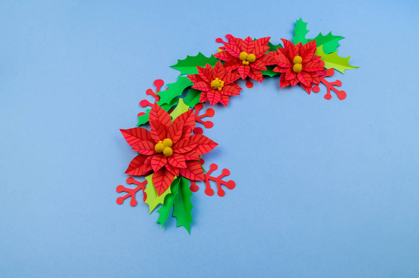 Christmas wreath of paper flowers poinsettia. Favorite hobby is manual work. Green holly and red berries. Blue background. - 写真・画像