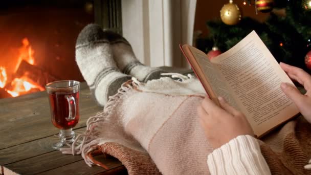 Zbliżenie 4k Zdjęcia młoda kobieta, leżąc z książki w salon z kominki i picie gorącej herbaty - Materiał filmowy, wideo
