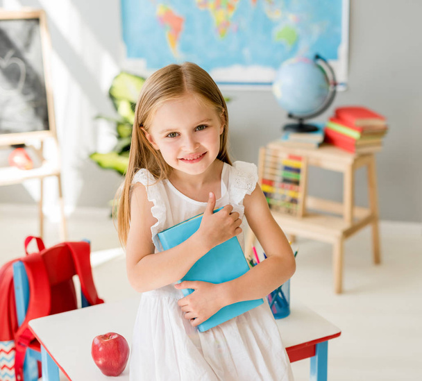 Little smiling blond girl holding blue book in the school class - Zdjęcie, obraz