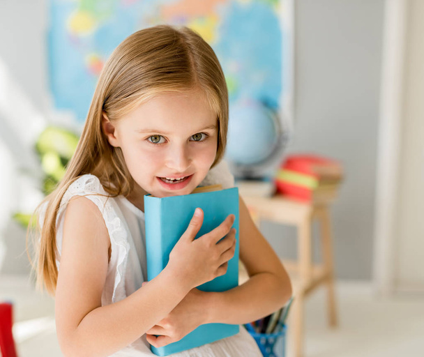 Little smiling blond girl holding blue book in the school class - Foto, imagen