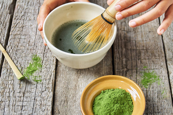 Matcha green tea accessoires on the rough wooden boards with girls hands preparing matcha tea in a clay bowl - Фото, изображение
