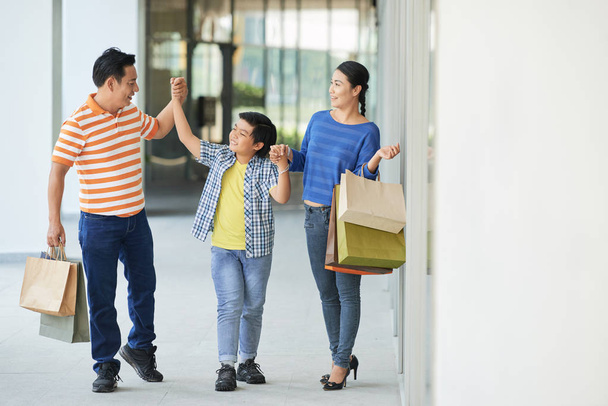 Amare i genitori con le borse della spesa in mano divertirsi con il loro piccolo figlio mentre si cammina lungo il corridoio del moderno centro commerciale
 - Foto, immagini