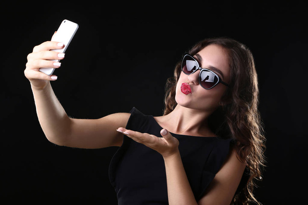 Young woman sending kiss and making selfie on black background - Φωτογραφία, εικόνα