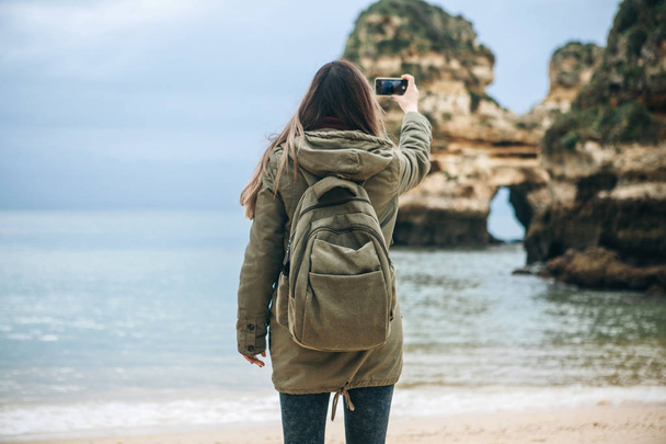 Girl tourist with a backpack photographing a beautiful landscape standing on the Atlantic coast - Photo, Image