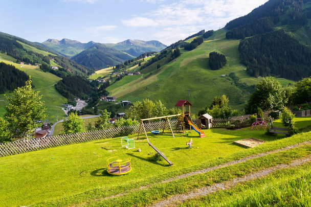 Hermoso parque infantil con columpio, tobogán, rotonda, caja de arena, gol de fútbol y casa de juegos en una colina con las montañas de los Alpes en el fondo, Saalbach Hinterglemm, Austria, día de verano soleado
 - Foto, Imagen
