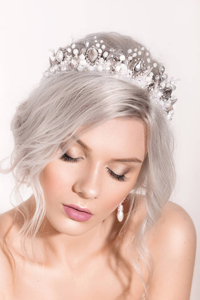 Beautiful young blond woman posing in crown and earrings in studio - 写真・画像