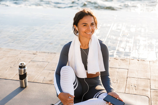 Image of young fitness smiling woman outdoors on the beach have a rest sitting with towel looking aside listening music with earphones. - Photo, image