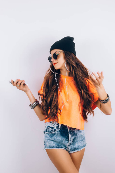 Young hipster beautiful woman in sunglasses wearing in black hat and orange T-shirt listening music in headphones near white wall, holding a cell phone in hand - Fotó, kép