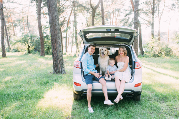 A cute young family has fun in the woods while sitting in the trunk of the car. - Фото, изображение