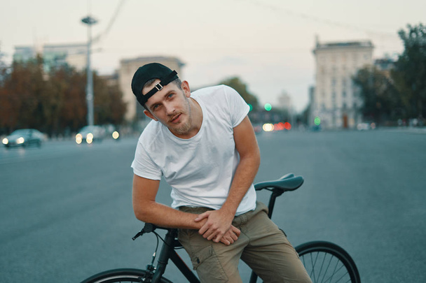Outdoor portrait of a modern young man in the street, sitting on bike. A Young athletic man wearing khaki pants, white T-shirt, looking to camera. - Photo, Image