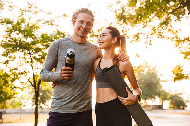 Picture of happy fitness sport loving couple friends in park outdoors holding bottle with water and fitness rug. - Foto, Bild