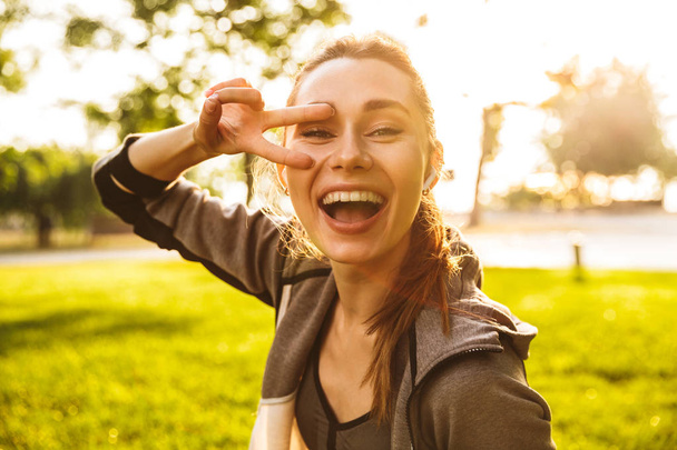 Photo of happy sporty woman 20s in sportswear wearing bluetooth earpod taking selfie with fingers while walking in green park - Fotografie, Obrázek