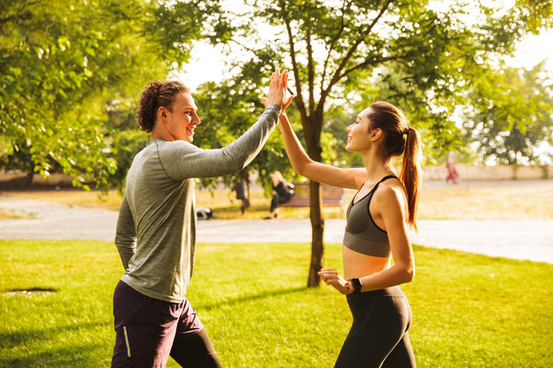 Photo of happy young man and woman 20s in tracksuits doing workout together in green park during sunny summer day - Fotó, kép