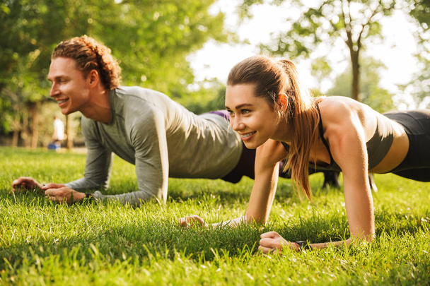 Photo of beautiful sporty couple man and woman 20s in sportswear doing plank together on grass while training in green park during sunny summer day - Foto, Bild