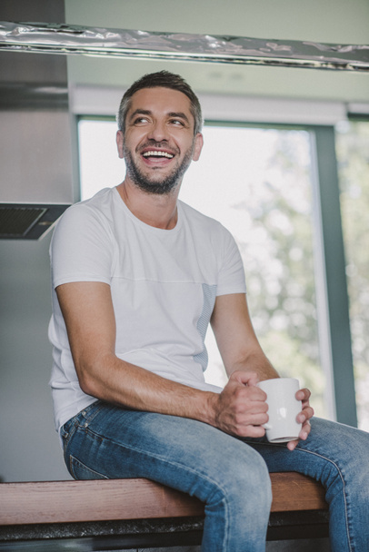 smiling handsome man sitting on kitchen counter with cup of tea in morning and looking away - Φωτογραφία, εικόνα