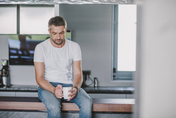 handsome sad man sitting on kitchen counter with cup of tea in morning - Фото, зображення