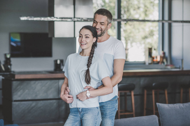 happy man hugging girlfriend from behind in kitchen at home - Photo, Image