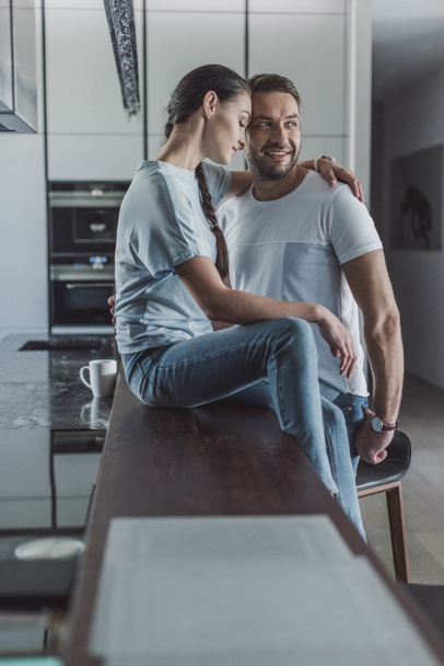 side view of attractive woman sitting on kitchen counter and embracing boyfriend at home - Fotografie, Obrázek