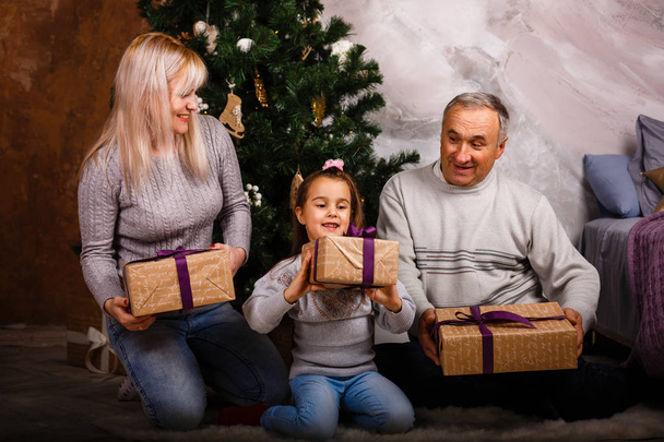 Little girl having fun with mom and grandfather sitting by Christmas tree at home  - Photo, Image