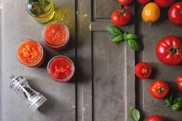 Variety of three homemade tomato sauces in glass jars with ingredients above. Different kinds of tomatoes, basil, olive oil, pepper, salt over old grey wooden background. Flat lay, space - Photo, Image