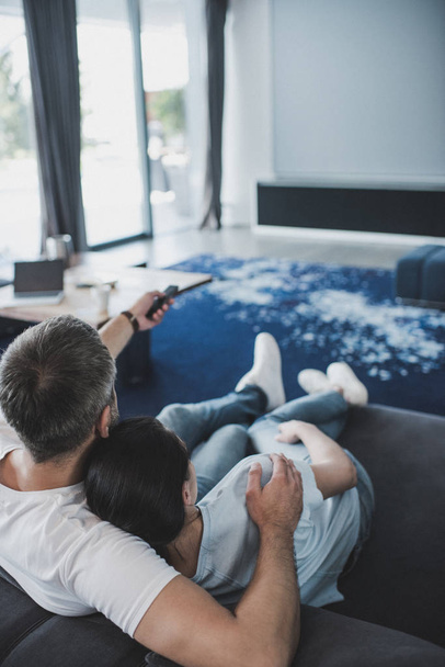 high angle view of adult man with remote controller embracing girlfriend and watching tv on sofa at home - Foto, Imagem