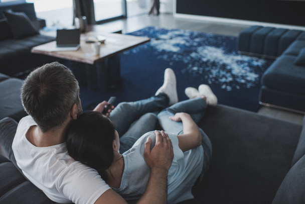 selective focus of adult man with remote controller embracing girlfriend and watching tv on sofa at home - Фото, изображение