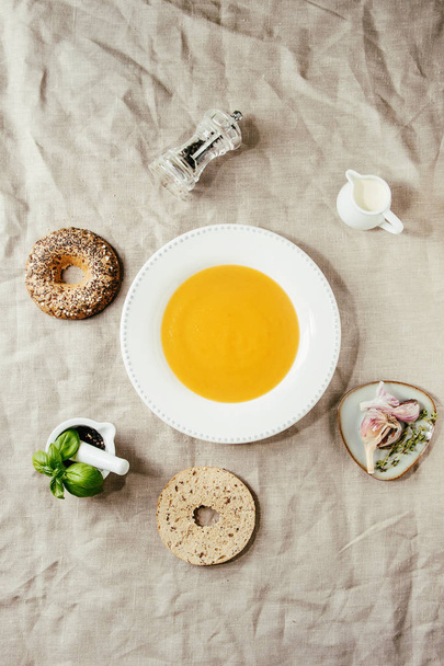 Plate of vegetarian pumpkin carrot soup served with with herbs, spoon, jug of cream, bagel bread, black pepper, garlic above over linen cloth as background. Flat lay, space - Photo, image