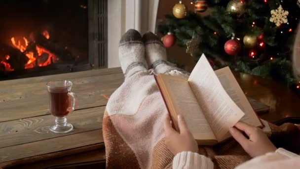 Vídeo em câmera lenta da jovem mulher lendo livro no sofá ao lado da lareira em chamas e bebendo chá na noite de Natal
 - Filmagem, Vídeo