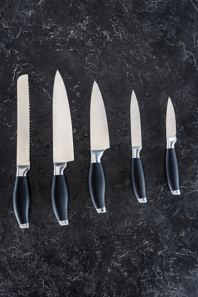 top view of various kitchen knives arranged on black marble surface   - Фото, изображение