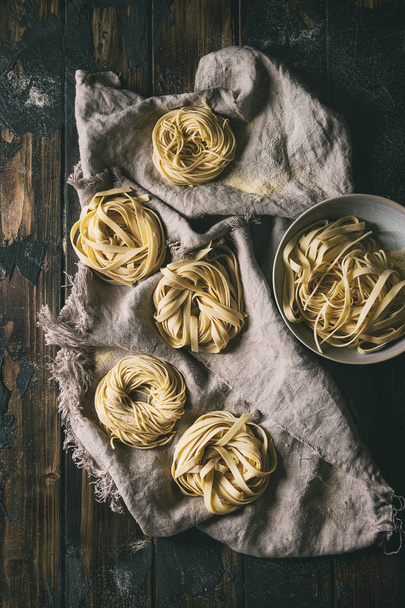 Variety of italian homemade raw uncooked pasta spaghetti and tagliatelle with semolina flour on linen cloth over dark plank texture wooden table. Flat lay, space. Dark rustic style - Foto, Imagem