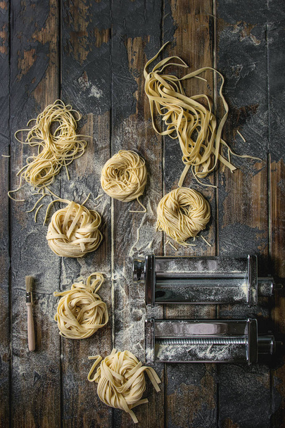 Variety of italian homemade raw uncooked pasta spaghetti and tagliatelle with pasta maker rolled machine and semolina flour over dark plank texture wooden table. Flat lay, space - Fotografie, Obrázek