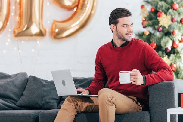 усміхнений чоловік з чашкою кави використовує ноутбук напередодні Різдва
 - Фото, зображення