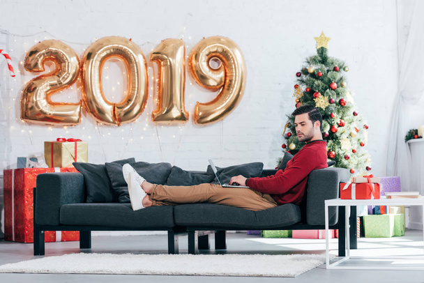 homme utilisant un ordinateur portable sur le canapé à la maison avec arbre de Noël et 2019 ballons dorés
 - Photo, image