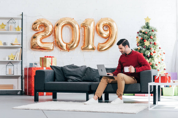 man with coffee cup using laptop at home with 2019 golden balloons and christmas tree behind - Foto, afbeelding