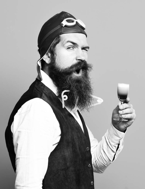 handsome bearded pilot or aviator man with long beard and mustache on funny face holding glass of alcoholic shot in vintage suede leather waistcoat with hat and glasses on blue studio background - Foto, Imagen