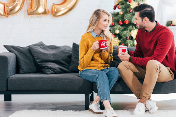 schönes Paar hält Tassen Tee und schaut sich zu Hause mit Weihnachtsbaum an - Foto, Bild
