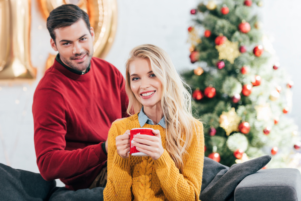 Улыбающаяся женщина держит чашку кофе, в то время как мужчина сидит дома с рождественской елкой
 - Фото, изображение