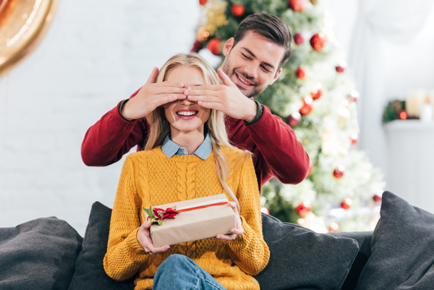 homme souriant faisant surprise avec boîte cadeau pour femme à la maison avec arbre de Noël
  - Photo, image
